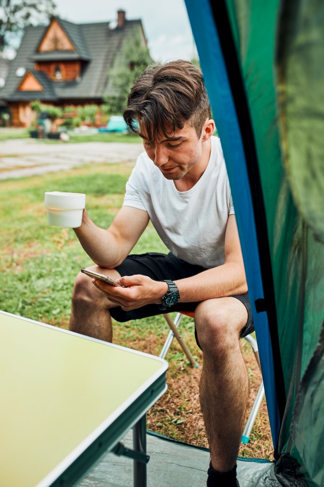 Young man drinking a coffee sitting at front of tent in the morning. Teenager enjoying free time during weekend trip in summer. Young man drinking a coffee sitting at front of tent in the morning