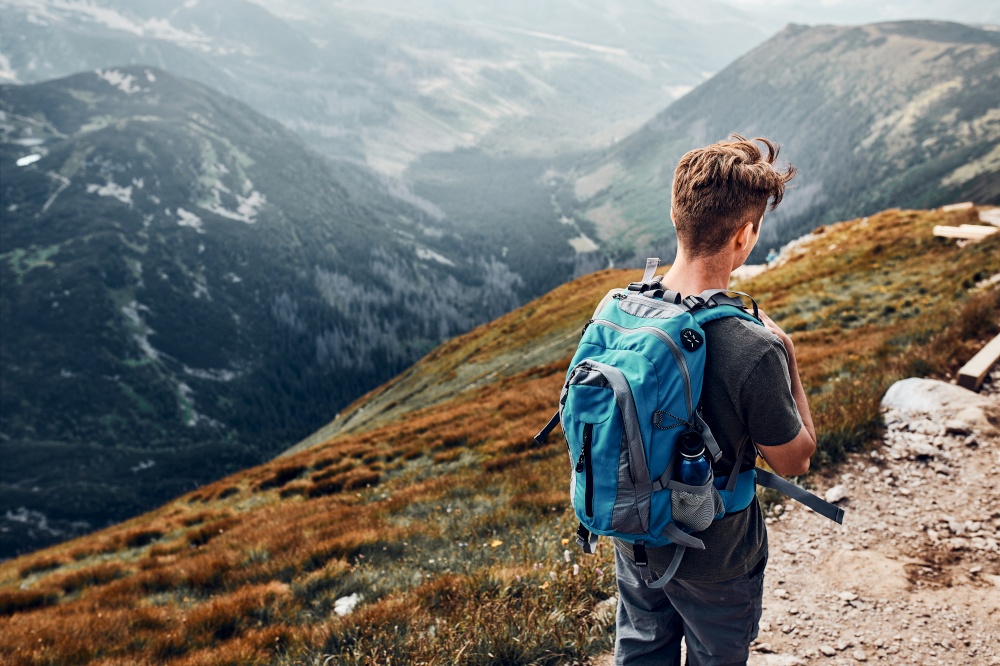 Young man with backpack hiking in a mountains, actively spending summer vacation. Rear view of teenager walking down from top of a hill along mountain path. Young man with backpack hiking in a mountains, actively spending summer vacation