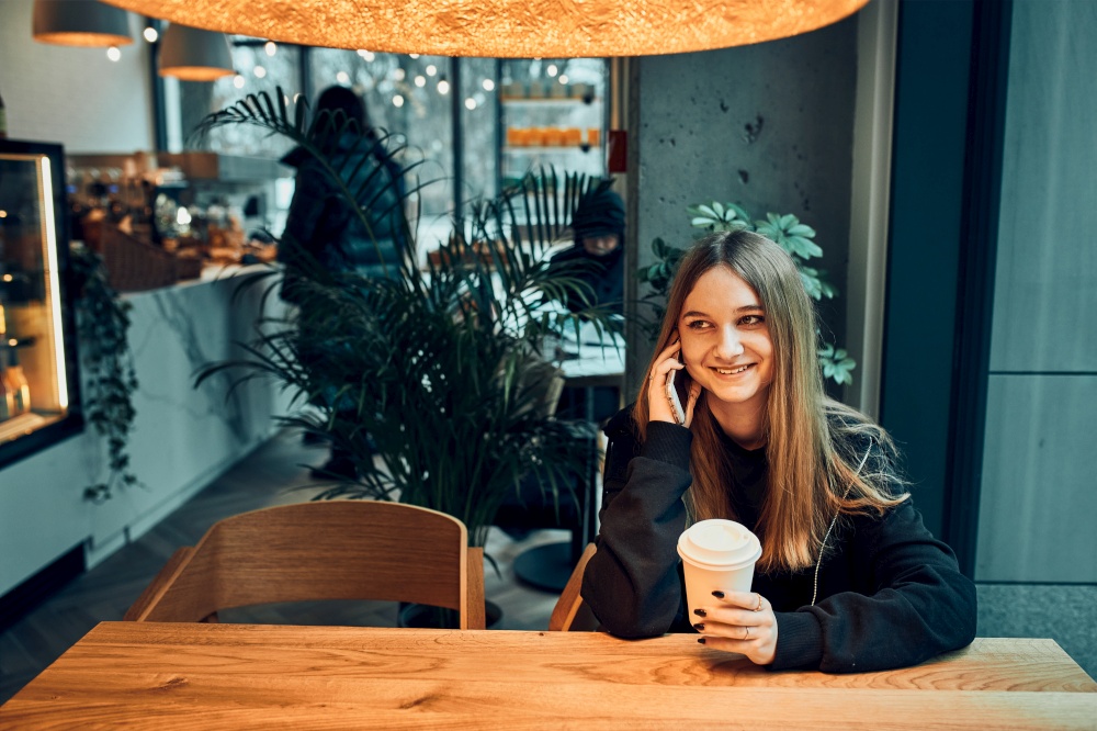 Young woman talking on the phone at cafe, having a pleasant phone call, answering call, chatting by mobile phone with friend while sitting in a cafe and drinking coffee. Girl relaxing in cafe