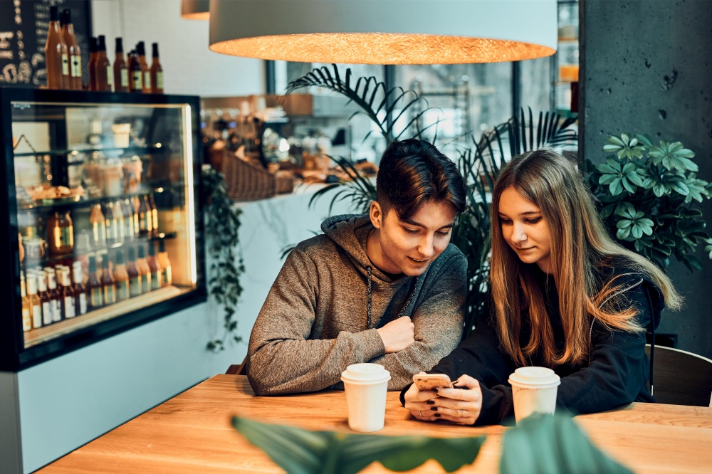 Couple relaxing in a cafe, using smartphone, having a chat, talking together, drinking coffee. Young man and woman having a break, spending time together