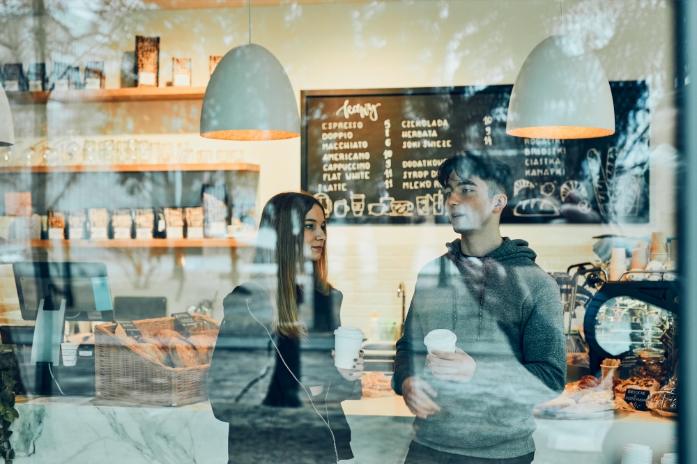 Friends having a chat, holding coffee cups, standing in a coffee shop and waiting for the their orders. Man and woman having a break drinking coffee in cafe. Interior view through the window