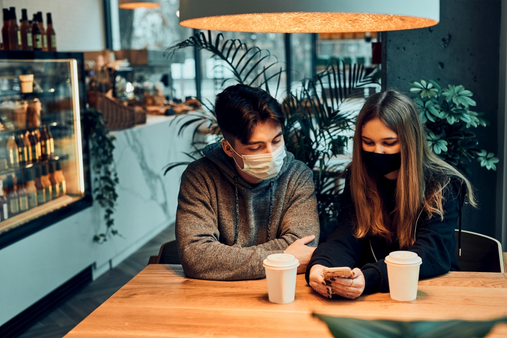 Couple relaxing in a cafe, using smartphone, having a chat, talking together, drinking coffee. Young man and woman having a break, spending time together. People wearing the face masks to avoid virus infection and to prevent the spread of disease in time of coronavirus. Couple relaxing in a cafe, using smartphone, having a chat, talking together, drinking coffee. Young man and woman having a break, spending time together. People wearing the face masks to avoid virus infection and to prevent the spread of disease in time