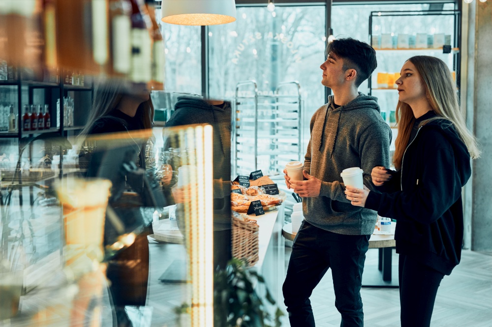 Friends doing shopping in a coffee shop. Young man and woman having chat while picking out the pastry, bakery&rsquo;s goods and hot drinks standing at counter in a coffee shop. People buying coffee and sweet snacks to go