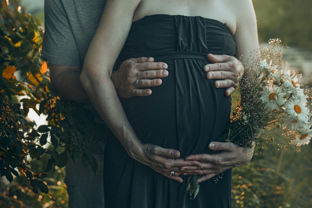 Pregnant woman and her husband hugging her tummy standing outdoors surrounded by nature. Pregnancy, expectation, motherhood concept