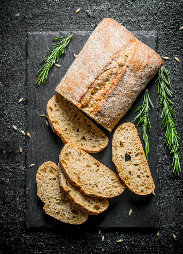 Sliced ciabatta bread on a stone Board with rosemary. On black rustic background. Sliced ciabatta bread on a stone Board with rosemary.