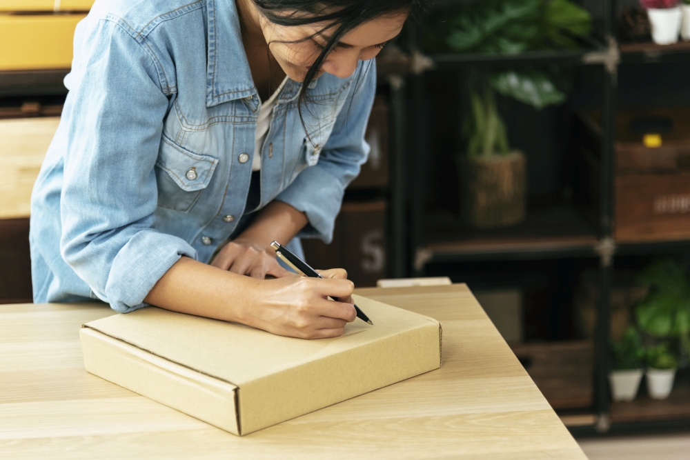 Woman packing box on table checking goods package delivery shipping to customer. Asian woman startup small business at home office desk. Entrepreneur asian woman packing product for delivery items