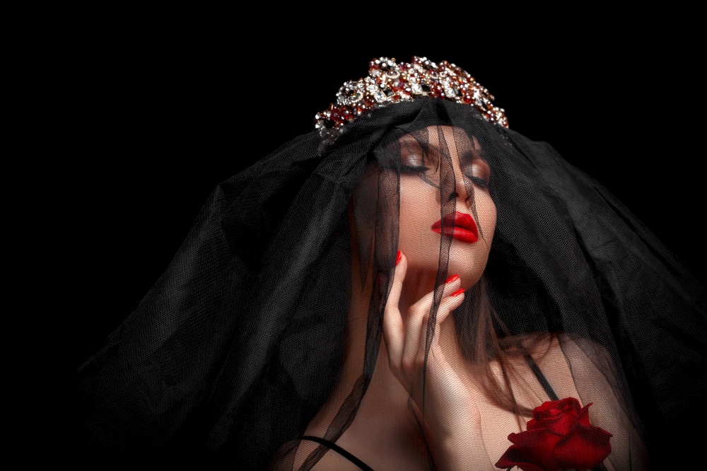 young beautiful girl in a black headband and black veil in a corset with a scarlet rose in her hands on a black isolated background./