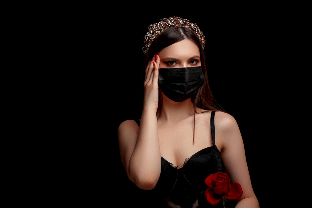 young beautiful girl with a headband on her head in a black medical mask with a scarlet rose in her hands on a black isolated background.