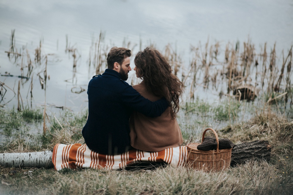 two caucasian lovers sitting on blanket by the lake. Young couple is hugging on autumn day outdoors. A bearded man and curly woman in love. Valentine&rsquo;s Day. Concept of love and family. back view.. two caucasian lovers sitting on blanket by the lake. Young couple is hugging on autumn day outdoors. A bearded man and curly woman in love. Valentine&rsquo;s Day. Concept of love and family. back view
