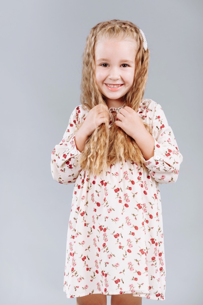 Portrait of a beautiful curly little girl in an elegant floral dress stands, laugh and looks into the camera in the studio on a gray background. Portrait of a beautiful curly little girl in an elegant floral dress stands, laugh and looks into the camera in the studio on a gray background.