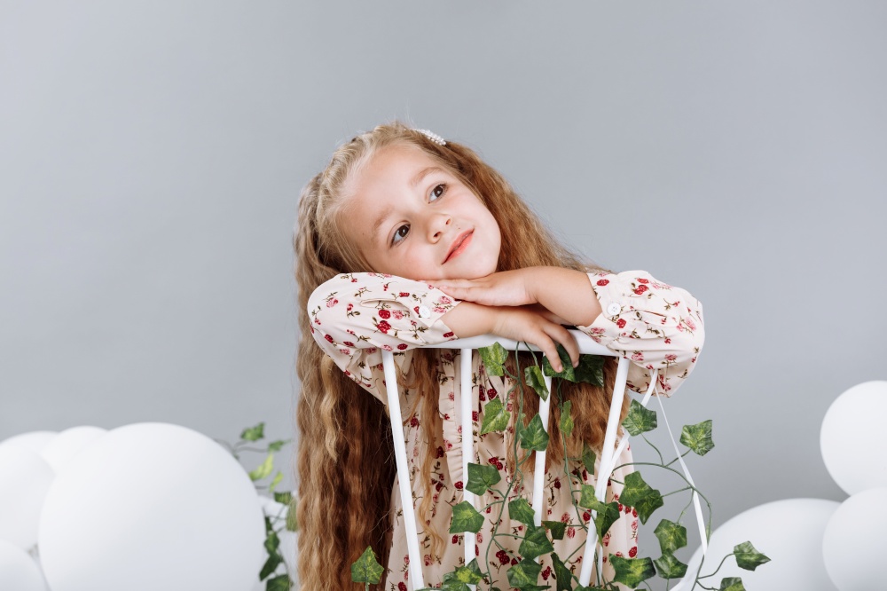 Cute little girl blonde smiling having fun in studio with white balloons and green. child in floral dress sitting on white chair and celebration easter. happy childhood.. Cute little girl blonde smiling having fun in studio with white balloons and green. child in floral dress sitting on white chair and celebration easter. happy childhood