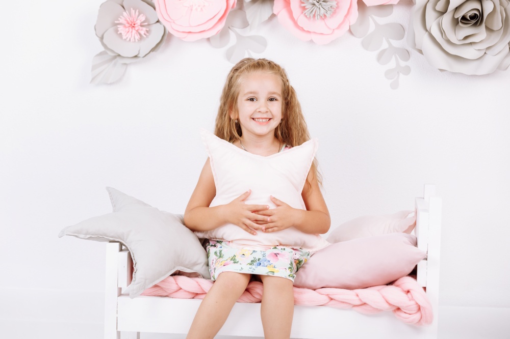 Bedtime. Little girl is sitting on bed with pink linen with pillow in the form of a star in her hands in white bedroom with handmade flowers on wall.. Bedtime. Little girl is sitting on bed with pink linen with pillow in the form of a star in her hands in white bedroom with handmade flowers on wall