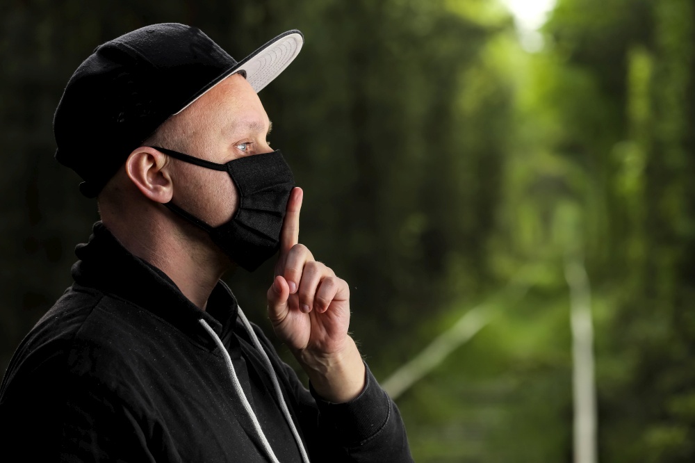 Young man in black protective antiviral mask with a finger to his lips in the summer park. The guy is resting outdoors in a mask made by his own hands. Virus protection. Quarantine measures.. Young man in black protective antiviral mask with a finger to his lips in the summer park. The guy is resting outdoors in a mask made by his own hands. Virus protection. Quarantine measures