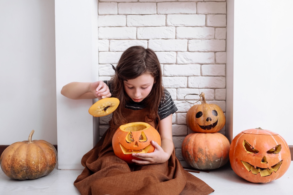 little girl with carving pumpkin on Halloween at home sitting next to fireplace in living room. Trick or treat. Child celebrating Halloween. little girl with carving pumpkin on Halloween at home sitting next to fireplace in living room. Trick or treat. Child celebrating Halloween.