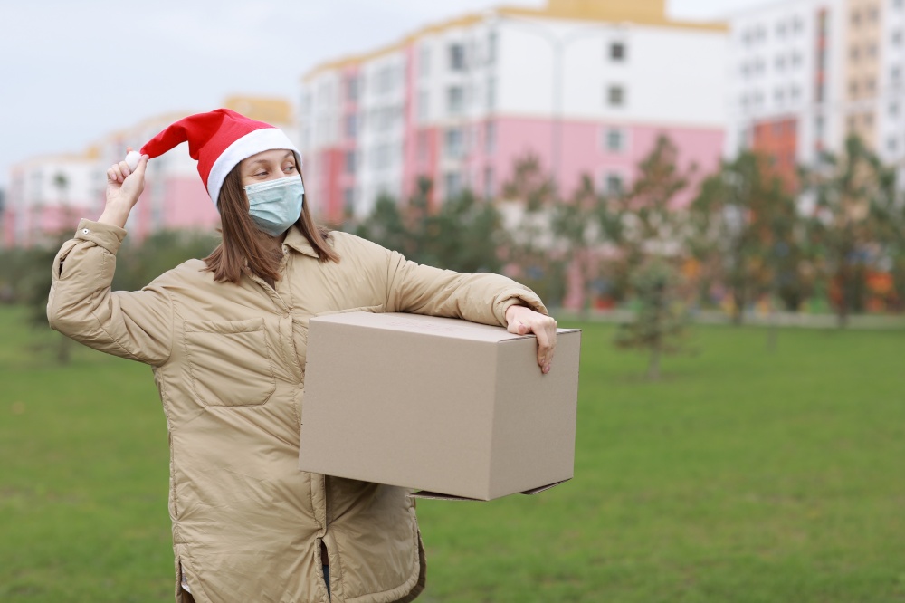 happy Delivery woman in a red Santa Claus hat and medical protective mask holds a big box outdoor. New Year&rsquo;s Christmas parcel in box, delivery online store in Quarantine time. Service coronavirus. happy Delivery woman in a red Santa Claus hat and medical protective mask holds a big box outdoor. New Year&rsquo;s Christmas parcel in box, delivery online store in Quarantine time. Service coronavirus.