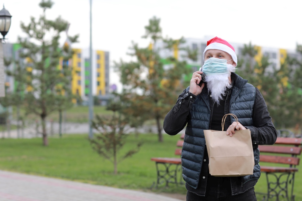 Delivery man in christmas uniform, protection mask holding delivery paper bag using smartphone to sending to customer. Take away food during quarantine in holiday time 2021. Delivery man in christmas uniform, protection mask holding delivery paper bag using smartphone to sending to customer. Take away food during quarantine in holiday time 2021.