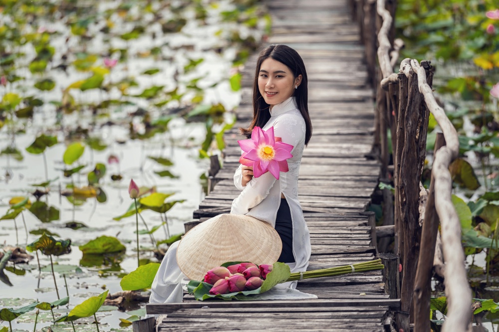 Portrait of beautiful vietnamese woman with traditional vietnam hat holding the pink lotus sitting on the wooden bridge in big lotus lake, vietnam, aisan or southeast asia travel concept
