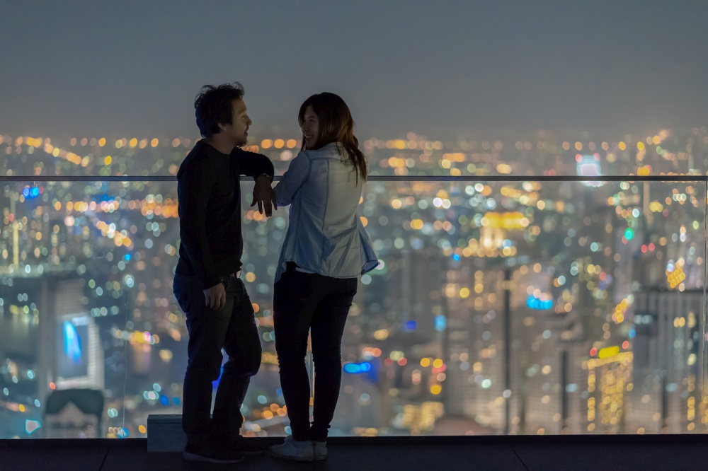 Romantic Couple standing on the rooftop from bangkok modern building at night time over the photo blurred of cityscape background, Lover and calentine holiday concept, low light