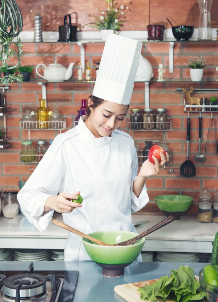 Female chef black hair white suit uniform apple and lemon on her hand preparing cooking in kitchen. Healthy food vegetable on woon plate, green bowl
