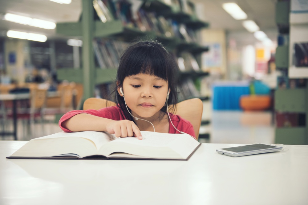 Cute little children girl studying read book at the library room and book on shelf background. children listen song music from smartphone on table