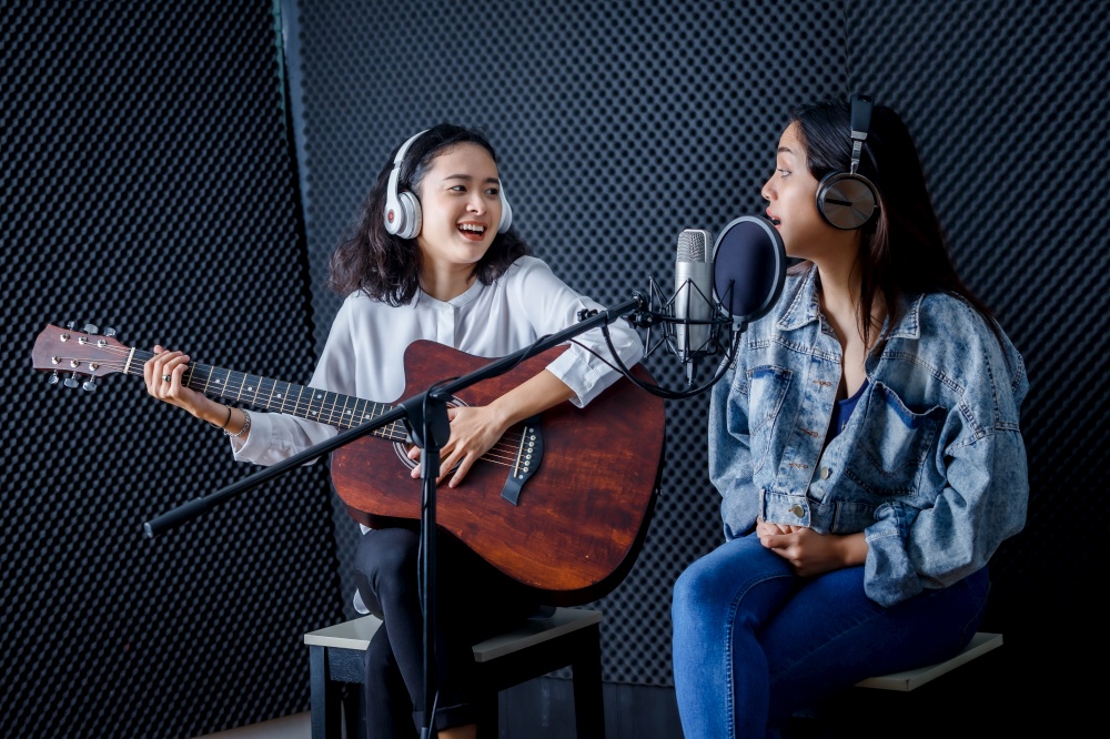 Happy cheerful pretty smiling of portrait Two young Asian woman vocalist Wearing Headphones with a guitar recording a song front of microphone in a professional studio