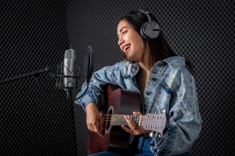 Happy cheerful pretty smiling of portrait a young Asian woman vocalist Wearing Headphones with a Guitar recording a song front of microphone in a professional studio