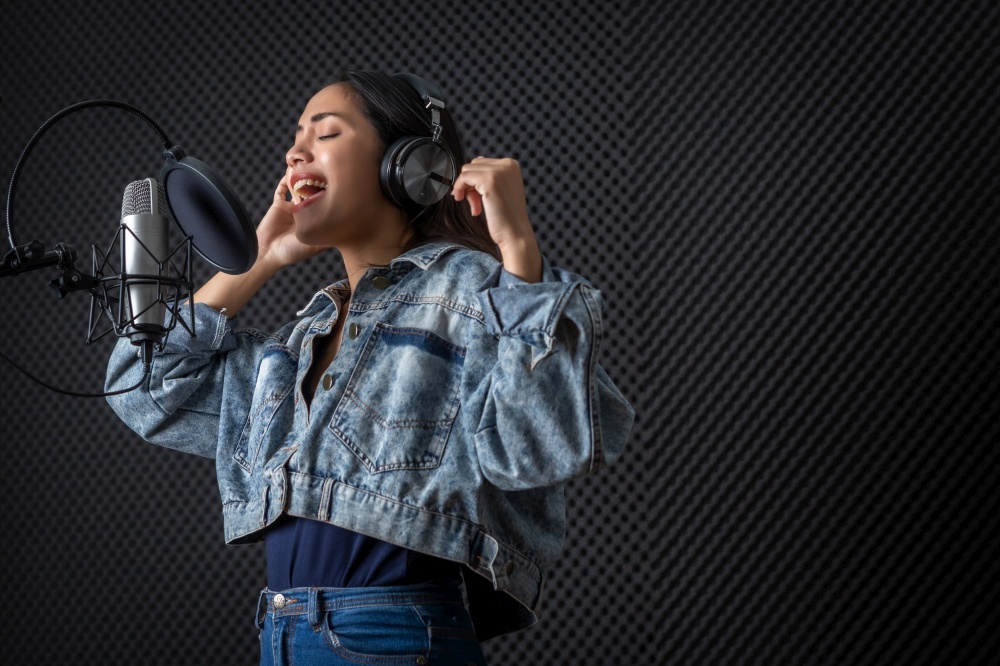 Happy cheerful pretty smiling of portrait of young Asian woman vocalist Wearing Headphones recording a song front of microphone in a professional studio