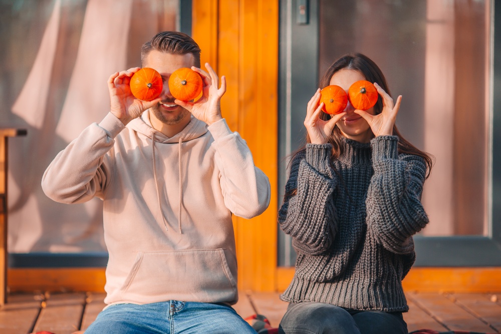 Young couple covered eyes with pumpkins at autumn warm day on the terrace of their house. Happy family of two on the terrace in autumn