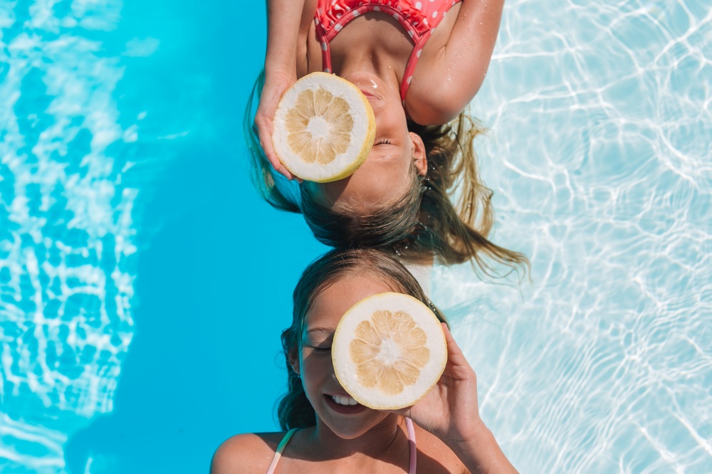 Portrait of girls in the pool look at camera. Little girls covering eyes with lemon halves near eyes on background swimming pool