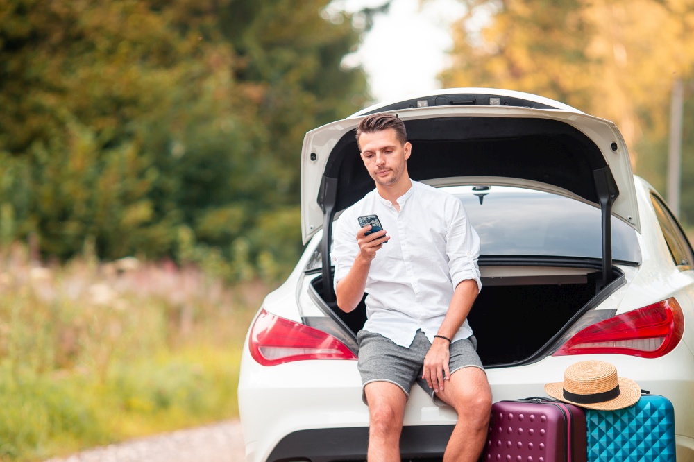 Man with smartphone sits in trunk of his car during his weekend. Young couple tourist enjoying on summer vacation