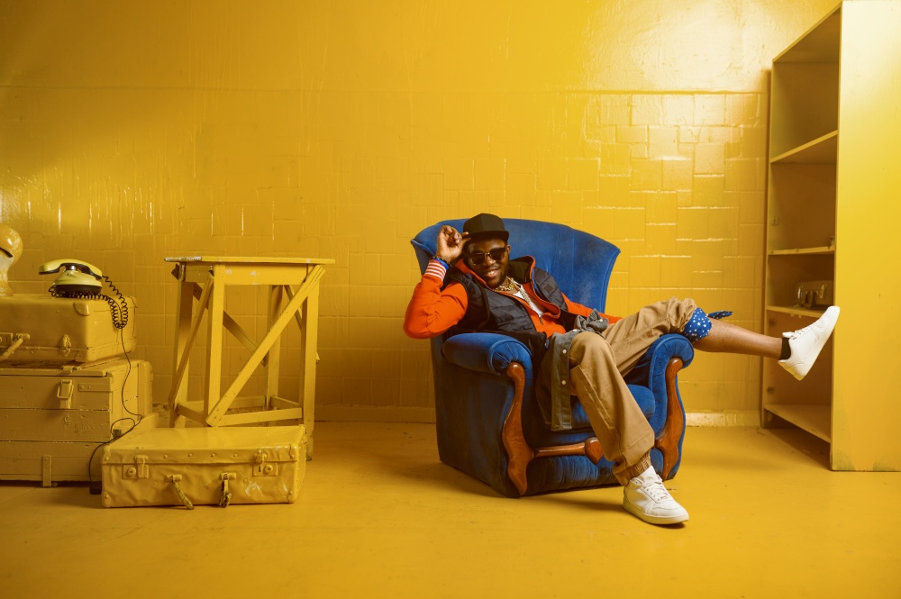 Smiling rapper posing in the chair in studio with yellow tones. Hip-hop performer, rap singer, break-dance performance. Rapper posing in chair in studio with yellow tones
