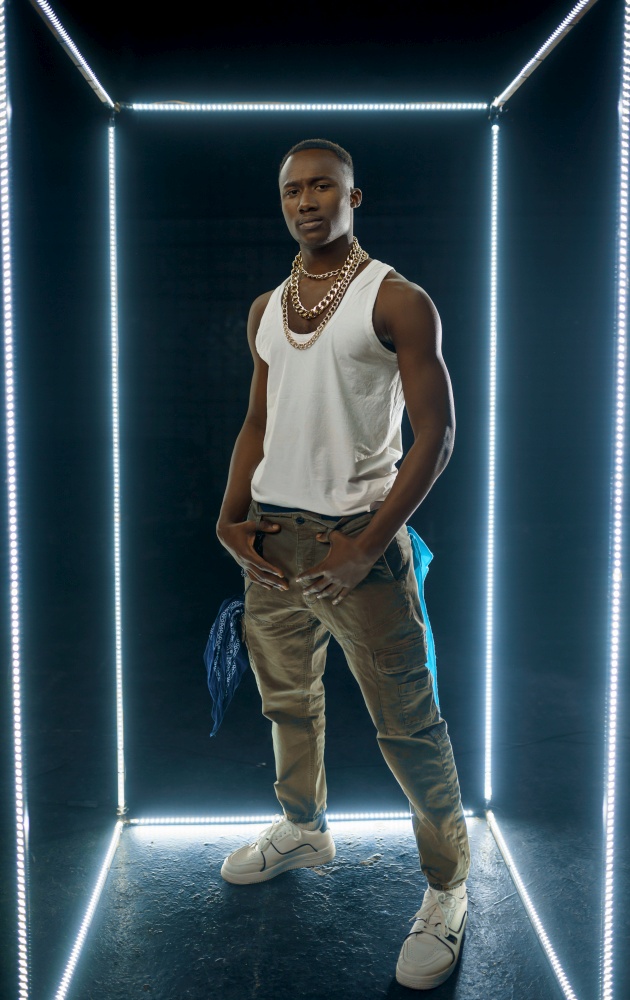 Serious rapper in gold jewelry poses in illuminated cube, dark background. Hip-hop performer, rap singer, break-dance performing, entertainment lifestyle, breakdancer. Rapper in gold jewelry poses in illuminated cube