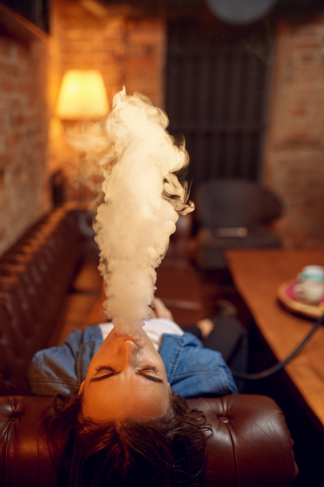 Young woman smokes on sofa in hookah bar, mesmerizing cloud. Shisha smoking, traditional bong culture, tobacco aroma for relaxation, rest with hooka. Young woman smokes on sofa in hookah bar