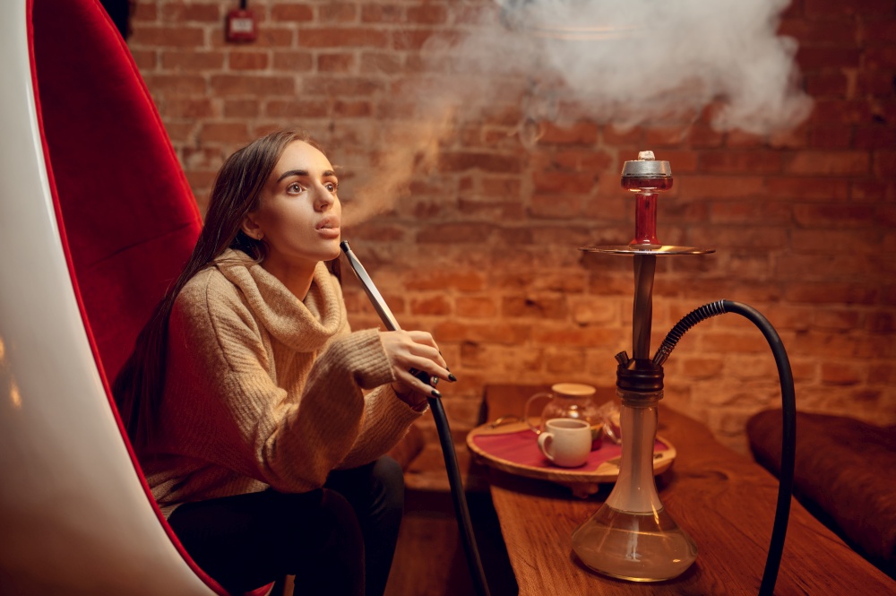 Young woman smokes hookah in bar, chill out. Shisha smoking, traditional bong culture, tobacco aroma for relaxation, rest with hooka. Young woman smokes hookah in bar, chill out