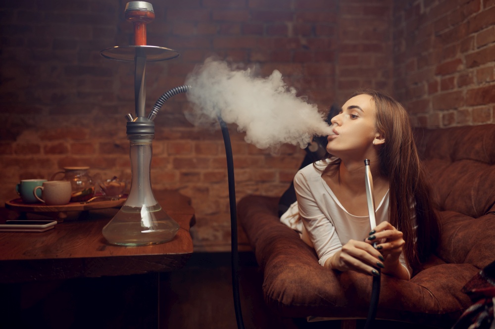 Young woman lying on sofa and smokes hookah in bar, chill out. Shisha smoking, traditional bong culture, tobacco aroma for relaxation, rest with hooka. Young woman lying on sofa and smokes hookah in bar
