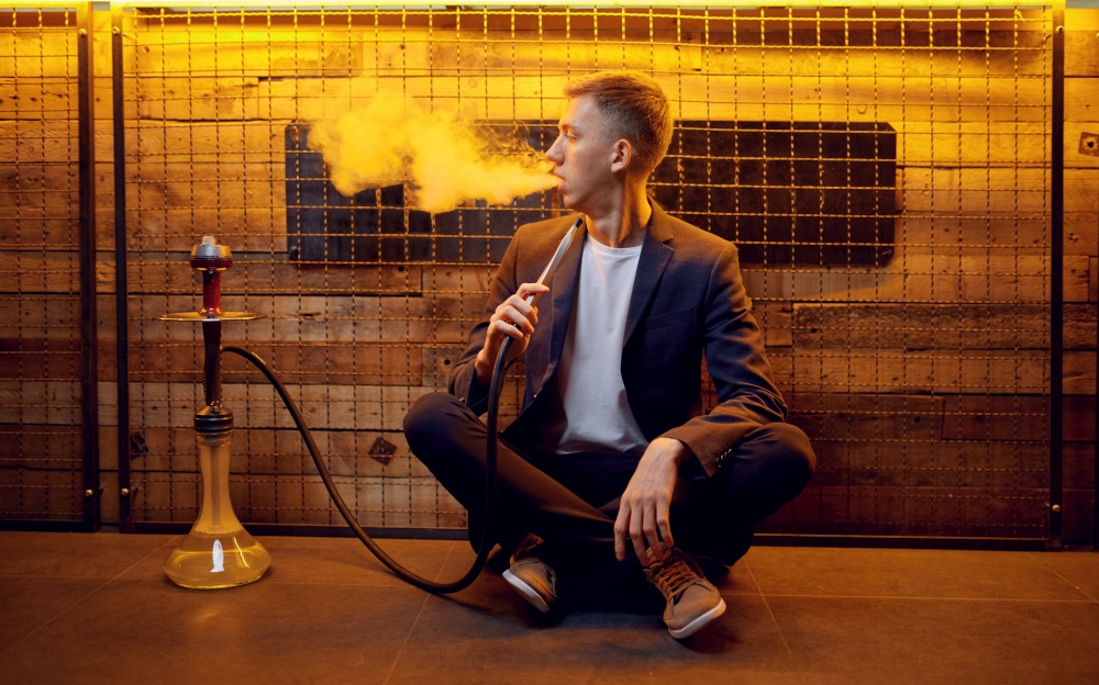 Young man smokes on the floor, hookah bar, hooka chill out. Shisha smoking, traditional bong culture, tobacco aroma for relaxation, rest with hooka. Young man smokes on the floor, hookah bar