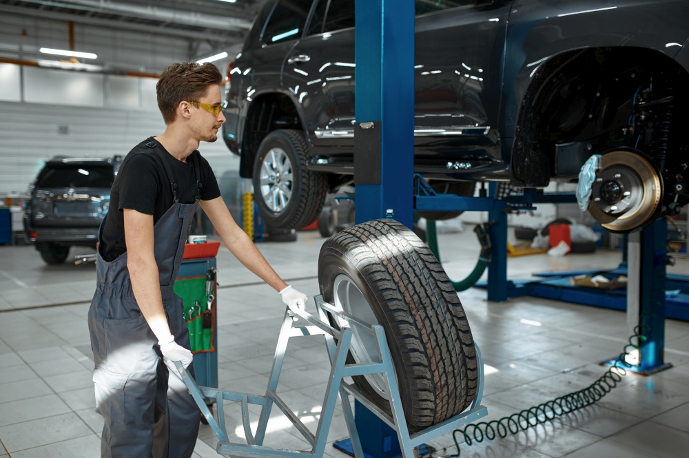 Male mechanic fixes problem with wheel, car service. Vehicle repairing garage, man in uniform, automobile station interior on background. Mechanic fixes problem with wheel, car service