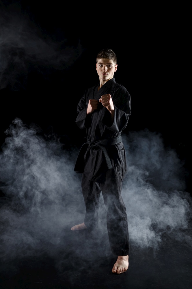Male karate fighter in black kimono, combat stance, dark background. Man on workout, martial arts, fighting competition. Male karate fighter in black kimono, combat stance