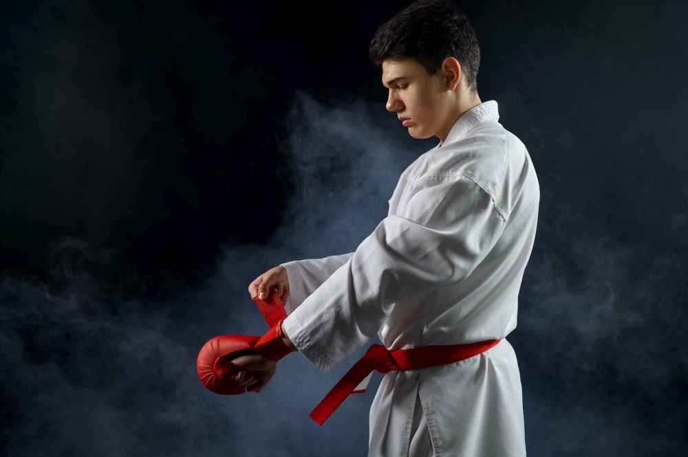 Male karate fighter in white kimono having red belt and gloves, combat stance, dark background. Karateka on workout, martial arts, training before fighting competition. Male karate fighter having red belt and gloves