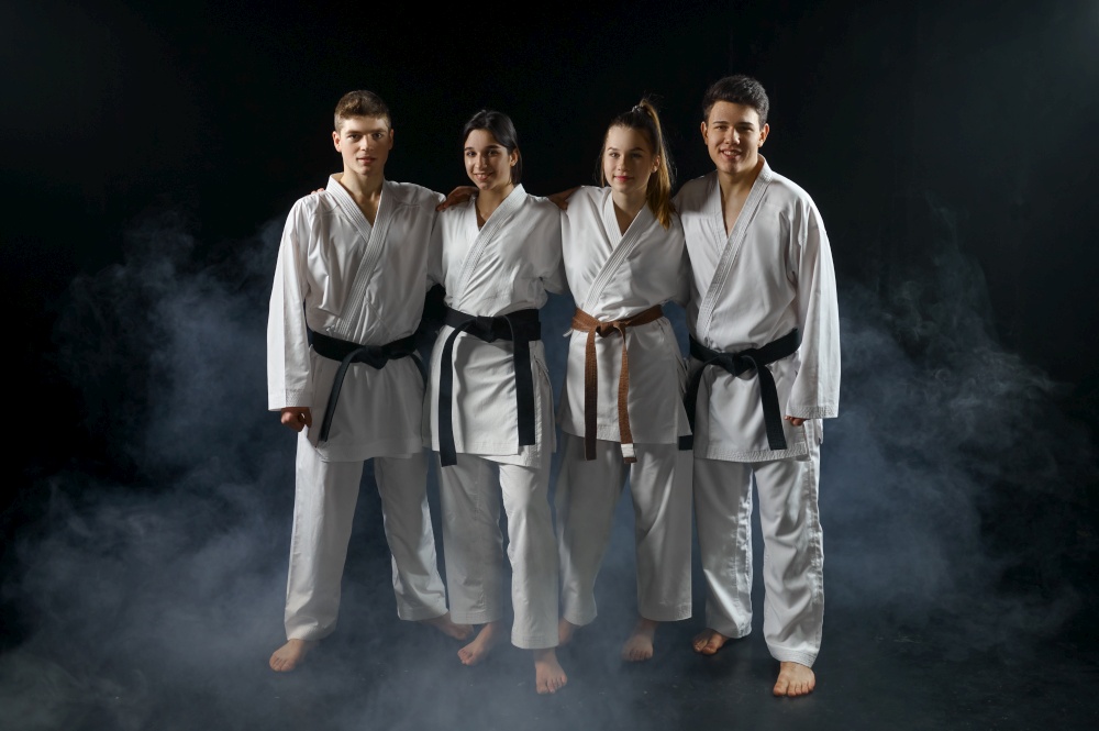 Four karate fighters in white kimono poses in a hug, group training, dark smoky background. Karatekas on workout, martial arts, fighting competition. Four karate fighters in white kimono poses in hug