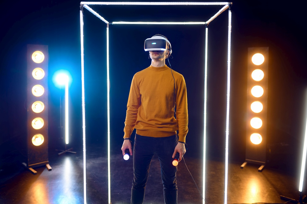 Gamer plays the game using virtual reality headset and gamepad in luminous cube. Dark playing club interior, spotlight on background, 3D VR technology. Gamer plays the game using virtual reality headset