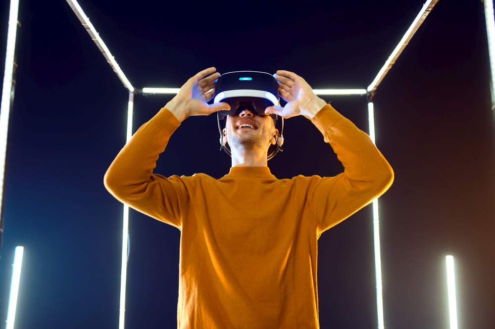 Male gamer plays the game using virtual reality headset and gamepad in luminous cube. Dark playing club interior, spotlight on background, VR technology with 3D vision. Male gamer using virtual reality headset