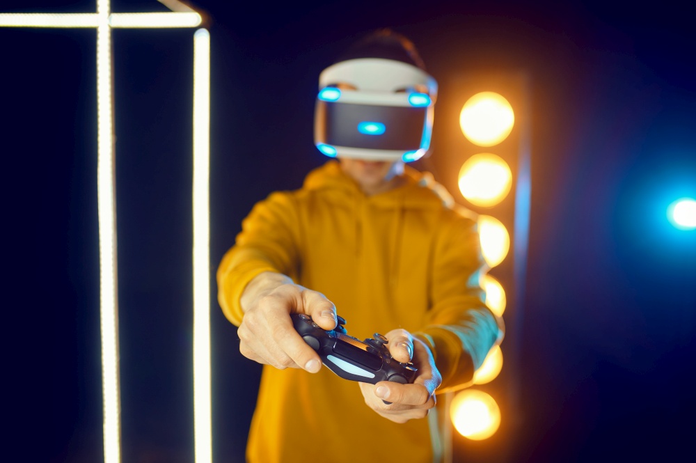 Man plays the game using virtual reality helmet and gamepad in luminous cube, front view. Dark playing club interior, spotlight on background, VR technology with 3D vision. Man using virtual reality helmet and gamepad