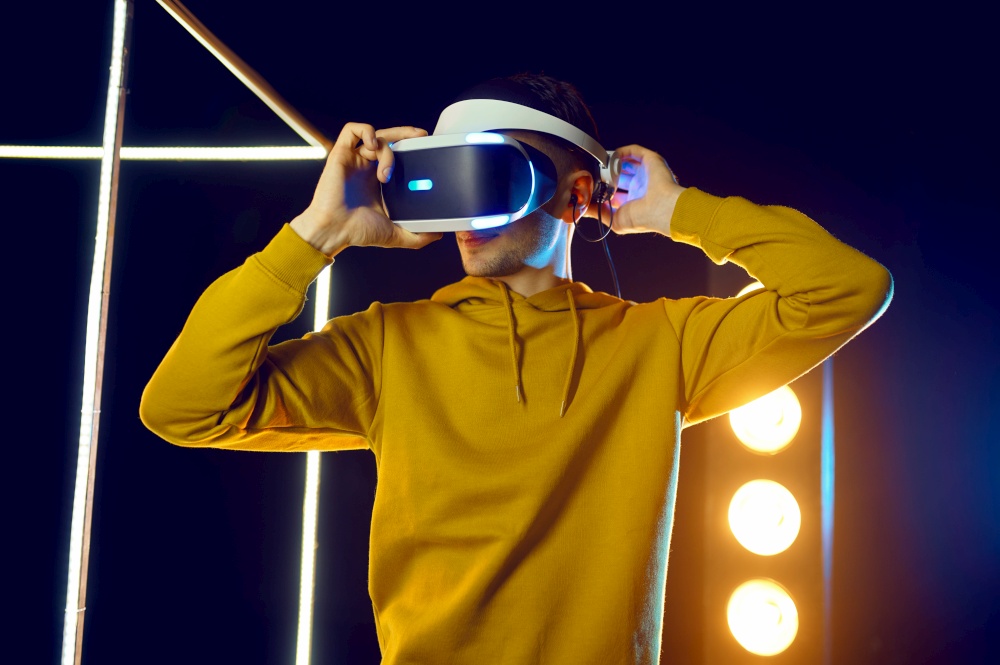 Young man plays the game using virtual reality headset and gamepad in luminous cube. Dark playing club interior, spotlight on background, VR technology with 3D vision. Man plays the game using virtual reality headset