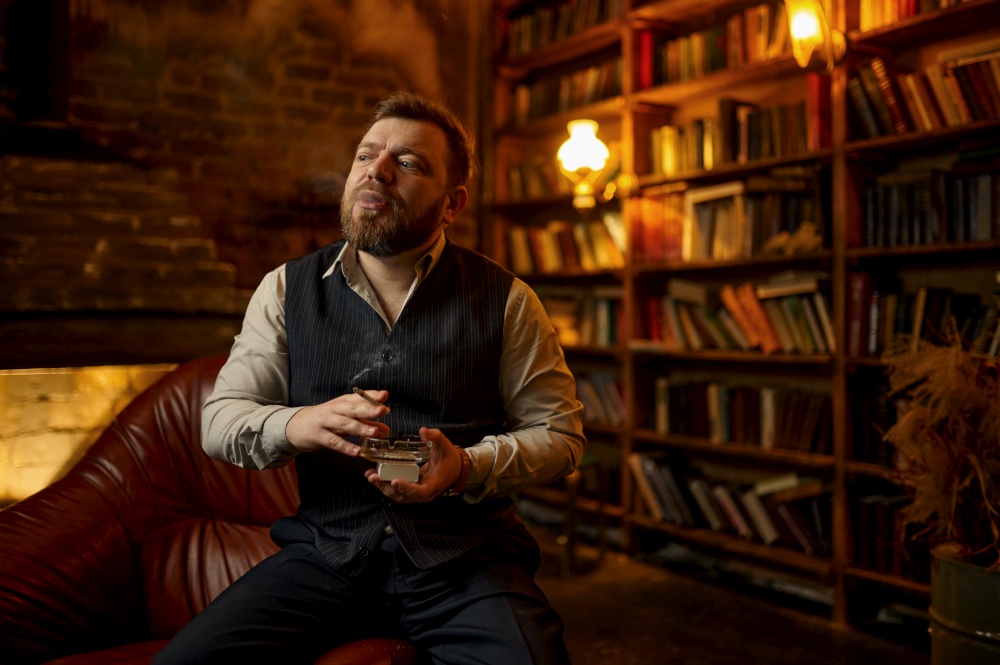 Bearded man holds ashtray and smokes cigarette, bookshelf and rich office interior on background. Tobacco smoking culture, specific flavor. Bearded man holds ashtray and smoking cigarette