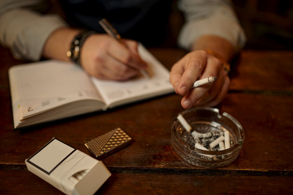 Man smokes cigarette and writes in notebook, wooden tabler on background. Tobacco smoking culture, specific flavor. Male smoker. Man smokes cigarette and writes in notebook