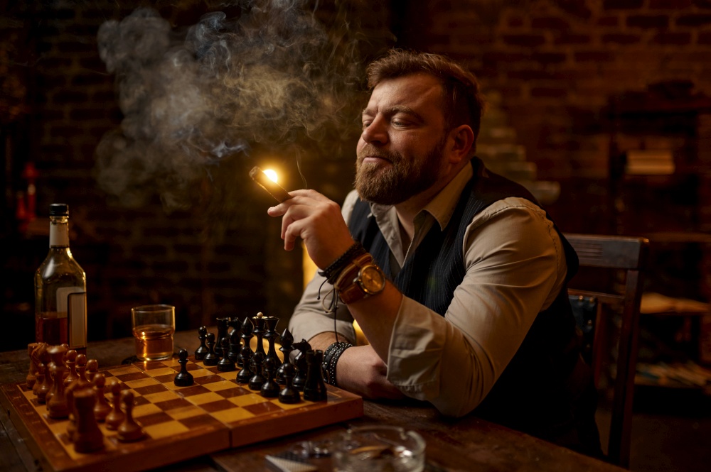Man smokes a cigar, drinks alcohol beverage and play chess, bookshelf and vintage office interior on background. Tobacco smoking culture, specific flavor. Male smoker leisures at the chessboard. Man smokes a cigar, smoker leisures at chessboard