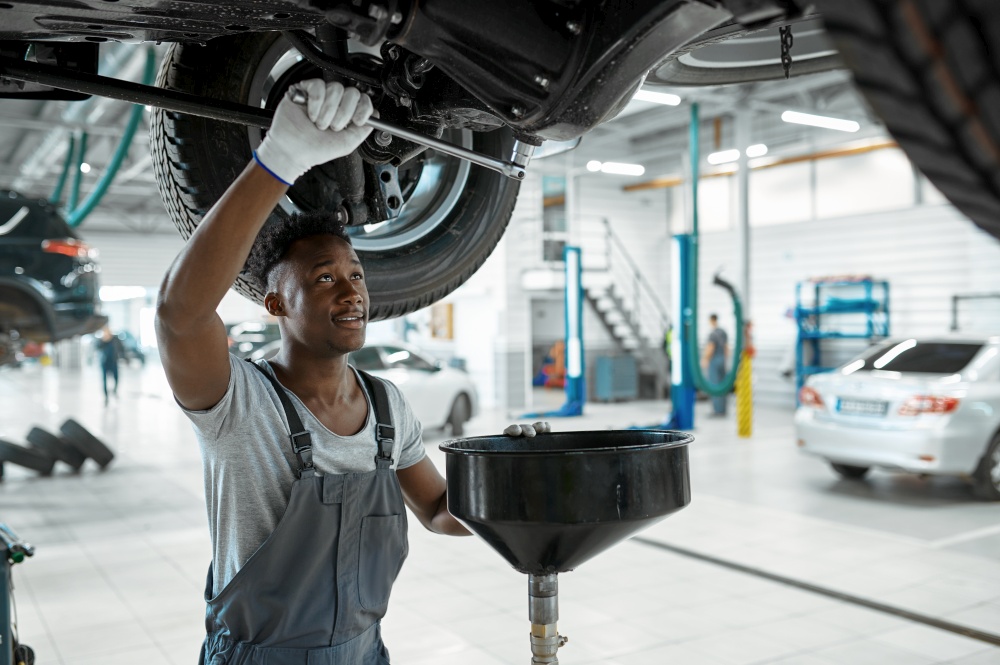 Male mechanic drains the oil, car service. Vehicle repairing garage, man in uniform, automobile station interior on background. Male mechanic drains the oil, car service