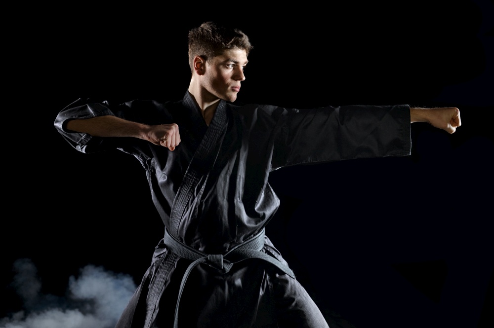 Male karate fighter in black kimono, combat stance, dark background. Man on workout, martial arts, fighting competition. Male karate fighter in black kimono, combat stance