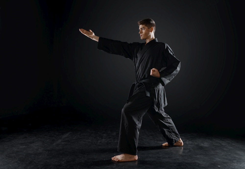 Male karateka, fighter practice in black kimono, combat stance, dark background. Man on workout, martial arts, training before fighting competition. Male karateka, fighter practice in black kimono